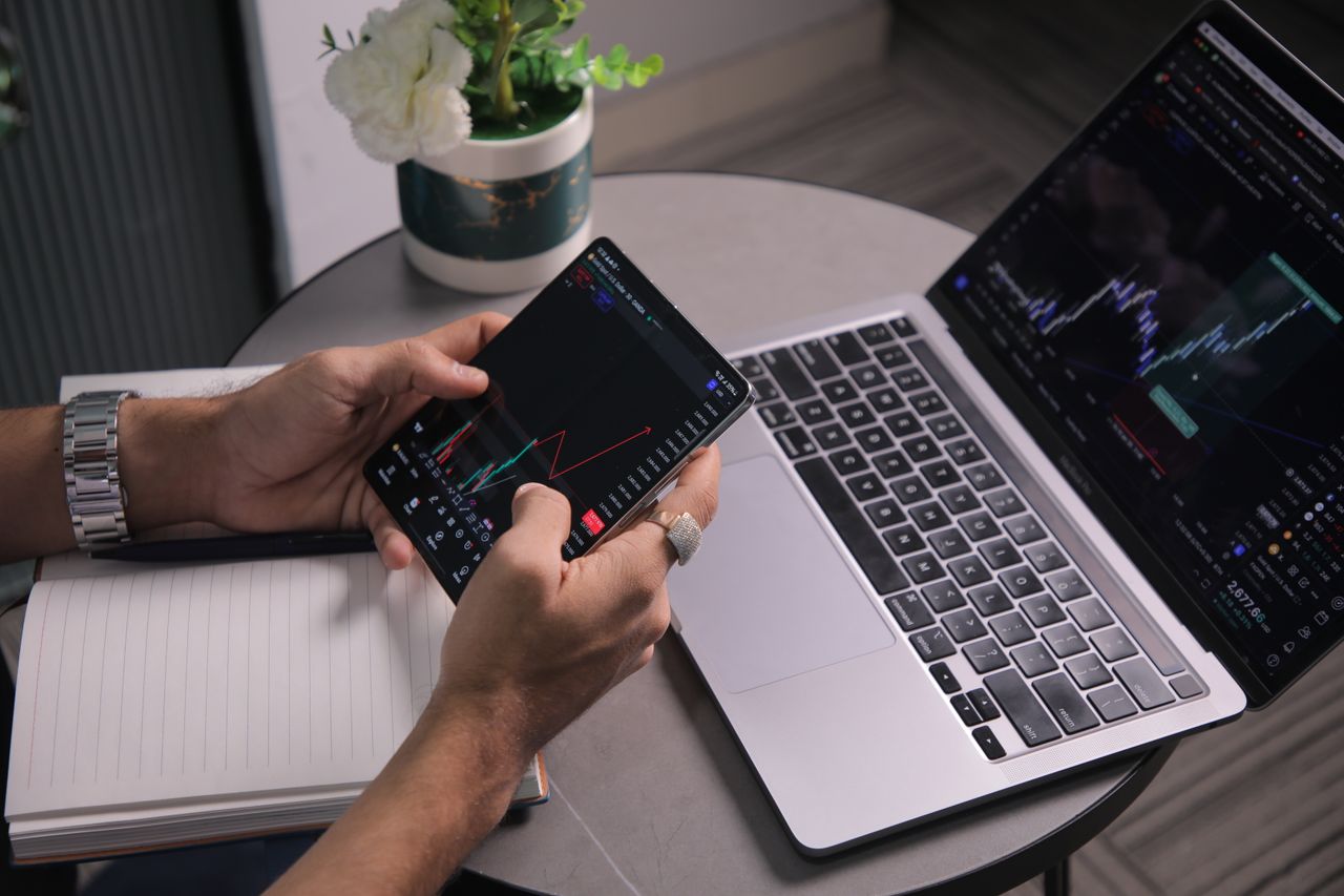 Un homme analyse les tendances boursières avec un ordinateur portable et une application de trading mobile dans un espace de travail moderne.